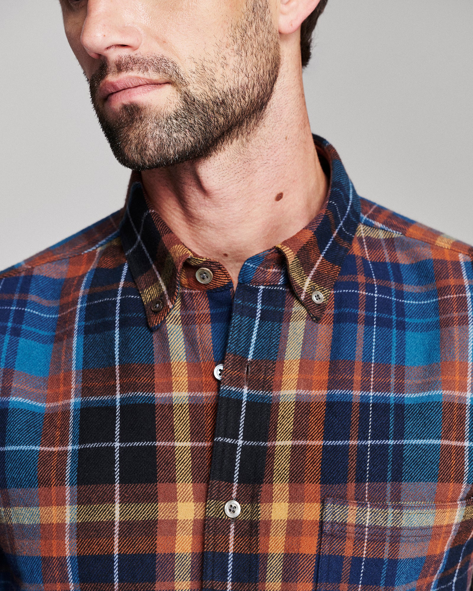 Close-up of a man wearing a button-down plaid shirt in blue, orange, black, and beige tones, showing the collar, buttons, and fabric texture against a plain light gray background.