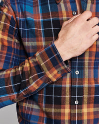 Close-up of a man wearing a long-sleeve plaid button-up shirt, showing his hand adjusting the collar and cuff. The shirt features a multi-colored plaid pattern with blue, orange, black, and yellow tones.