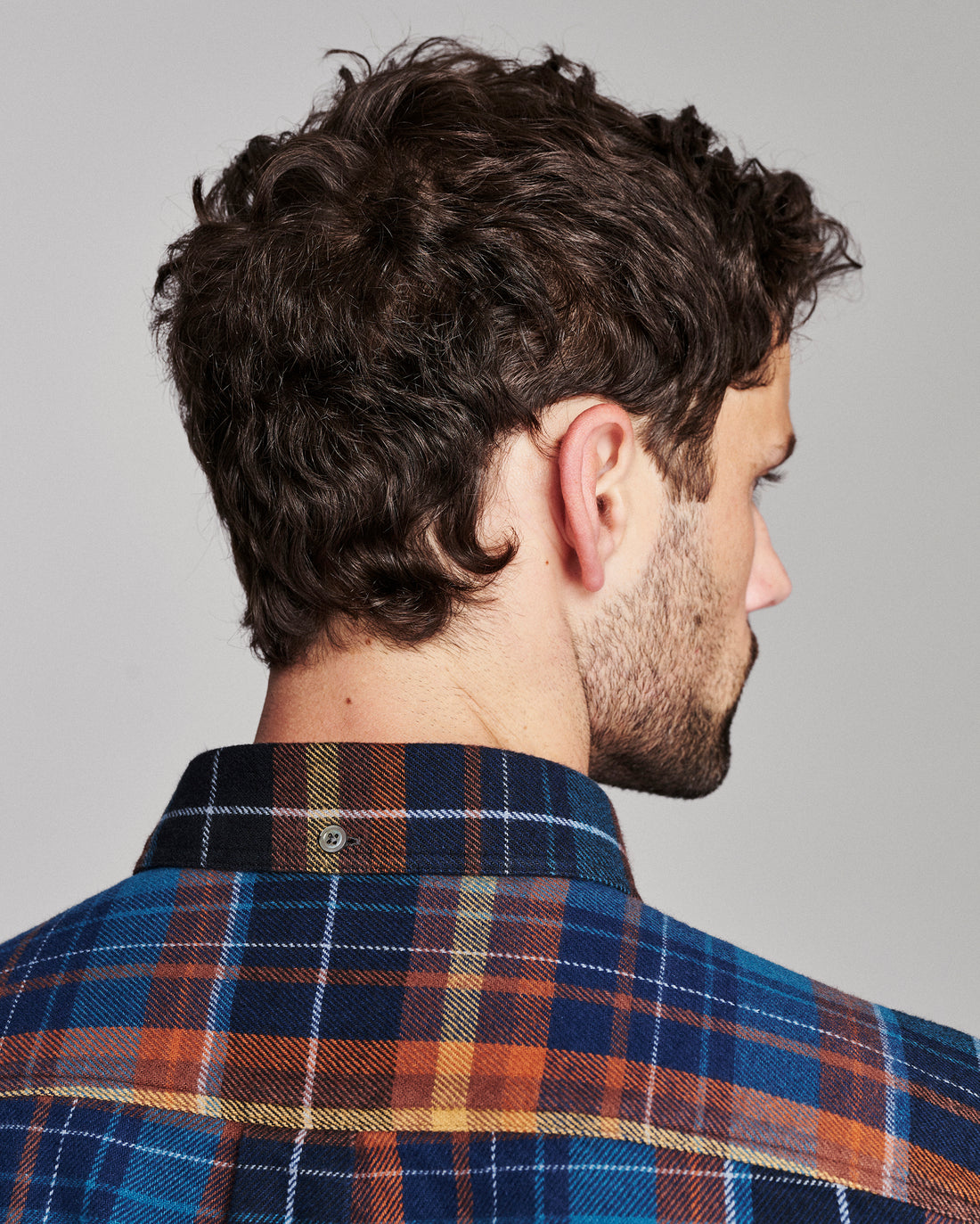 A man shown from the back and side profile wearing a blue, orange, and yellow plaid flannel shirt against a solid gray background.