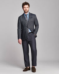 A man modeling a dark gray textured two-piece suit with a light blue dress shirt, patterned tie, and brown suede loafers, standing against a solid light gray background.
