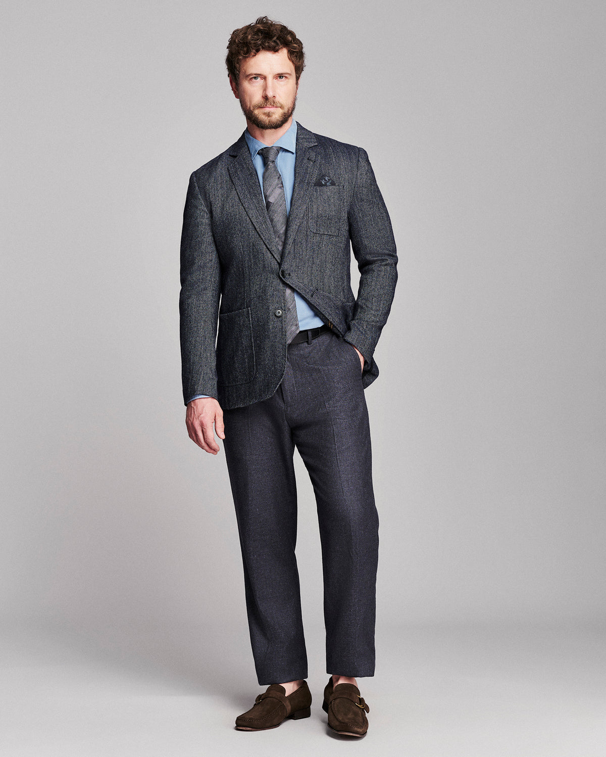 A man modeling a dark gray textured two-piece suit with a light blue dress shirt, patterned tie, and brown suede loafers, standing against a solid light gray background.