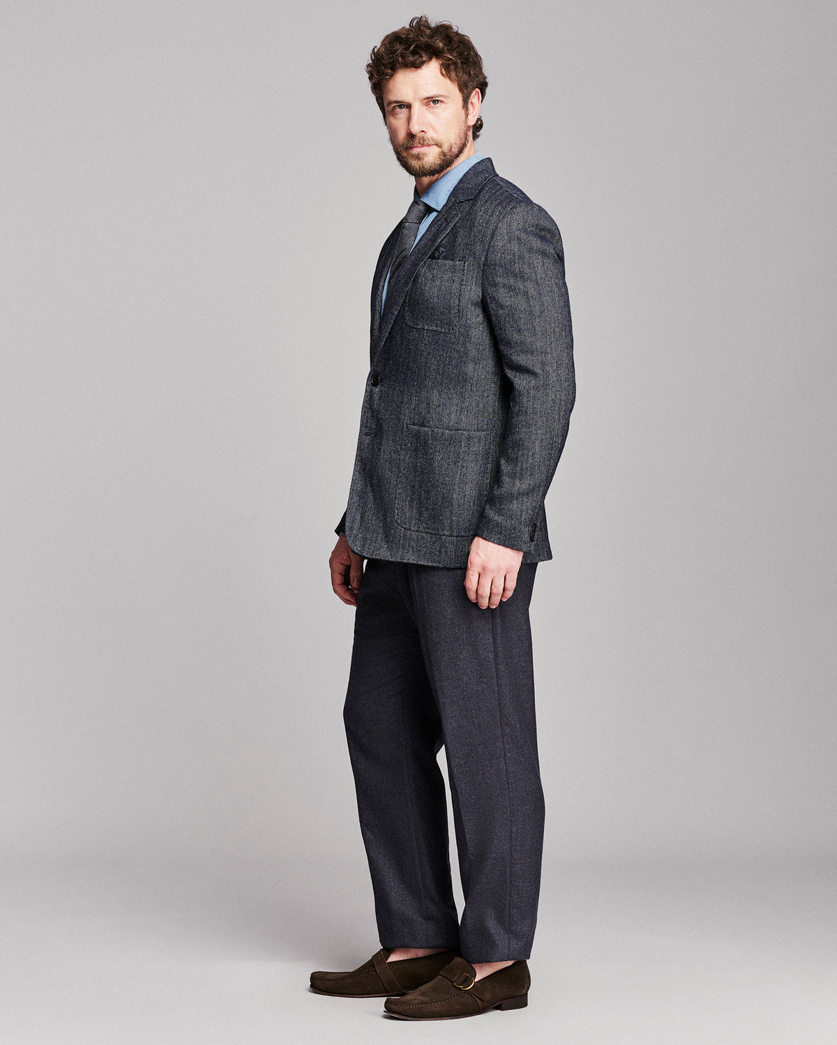 A man modeling a dark gray textured two-piece suit with a light blue dress shirt and brown loafers, standing against a solid gray studio background.