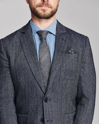 A close-up of a man wearing a textured charcoal gray suit jacket, a light blue dress shirt, a gray striped tie, and a matching pocket square, set against a solid gray background.