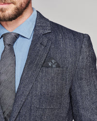 Close-up of a man wearing a dark gray herringbone tweed suit jacket, a light blue dress shirt, a gray tie, and a matching pocket square with a small embroidered logo.