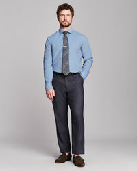 A man modeling a light blue dress shirt, a dark gray patterned tie, dark charcoal gray trousers, and brown leather loafers against a solid light gray studio background.