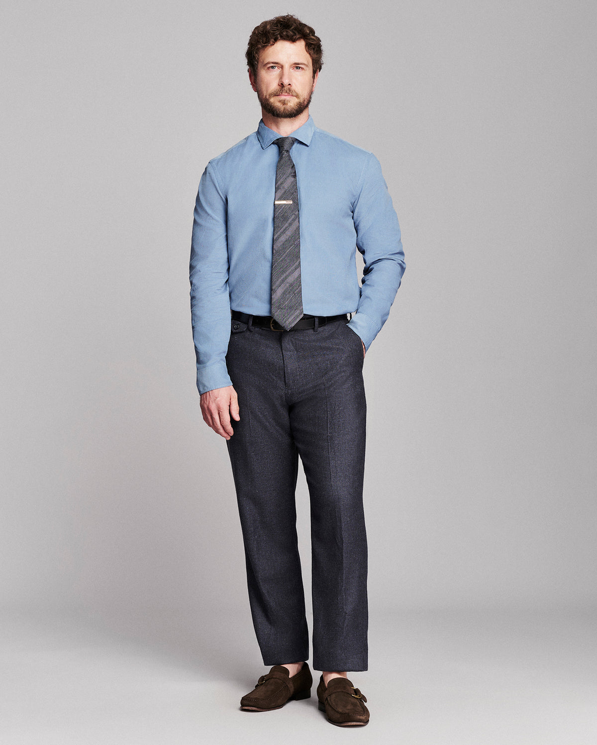 A man modeling a light blue dress shirt, a dark gray patterned tie, dark charcoal gray trousers, and brown leather loafers against a solid light gray studio background.