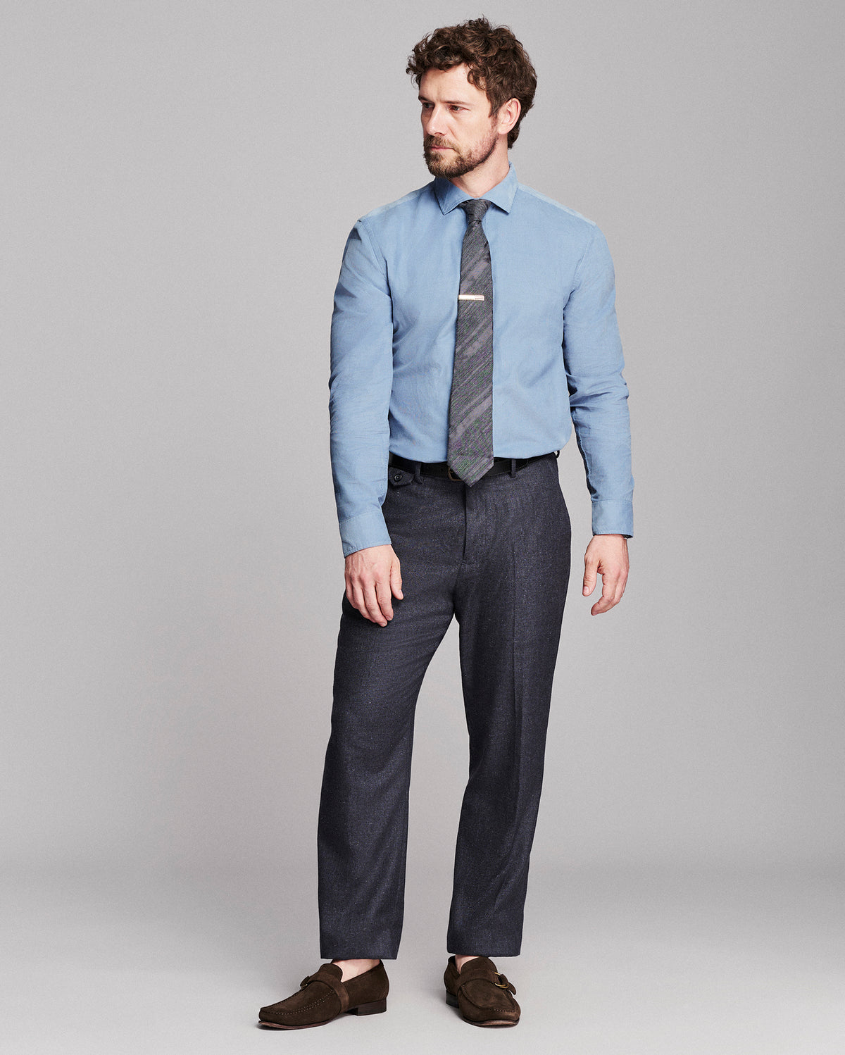 A man modeling a light blue dress shirt, a dark gray patterned tie, dark navy trousers, and brown suede loafers against a solid gray studio background.