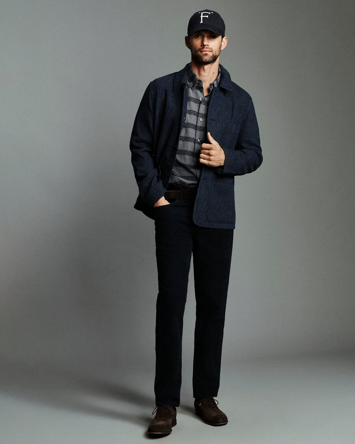 A man modeling a dark navy blue corduroy jacket over a gray and black plaid button-down shirt, paired with dark trousers, brown leather shoes, and a black baseball cap with a white 'F' logo, set against a solid gray studio background.