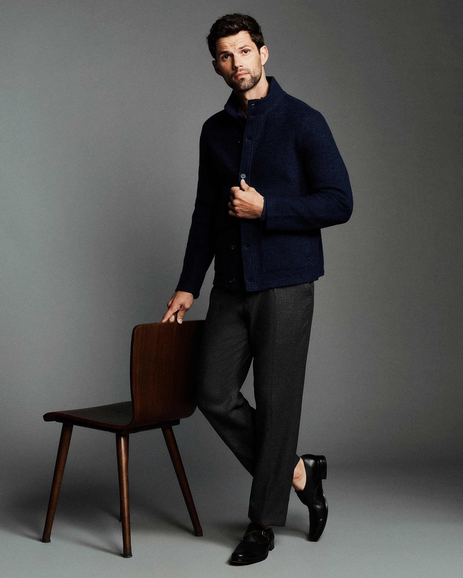 A man modeling a dark navy textured knit cardigan over a collared shirt, paired with charcoal gray trousers and black dress shoes, leaning against a wooden chair against a solid gray studio background.