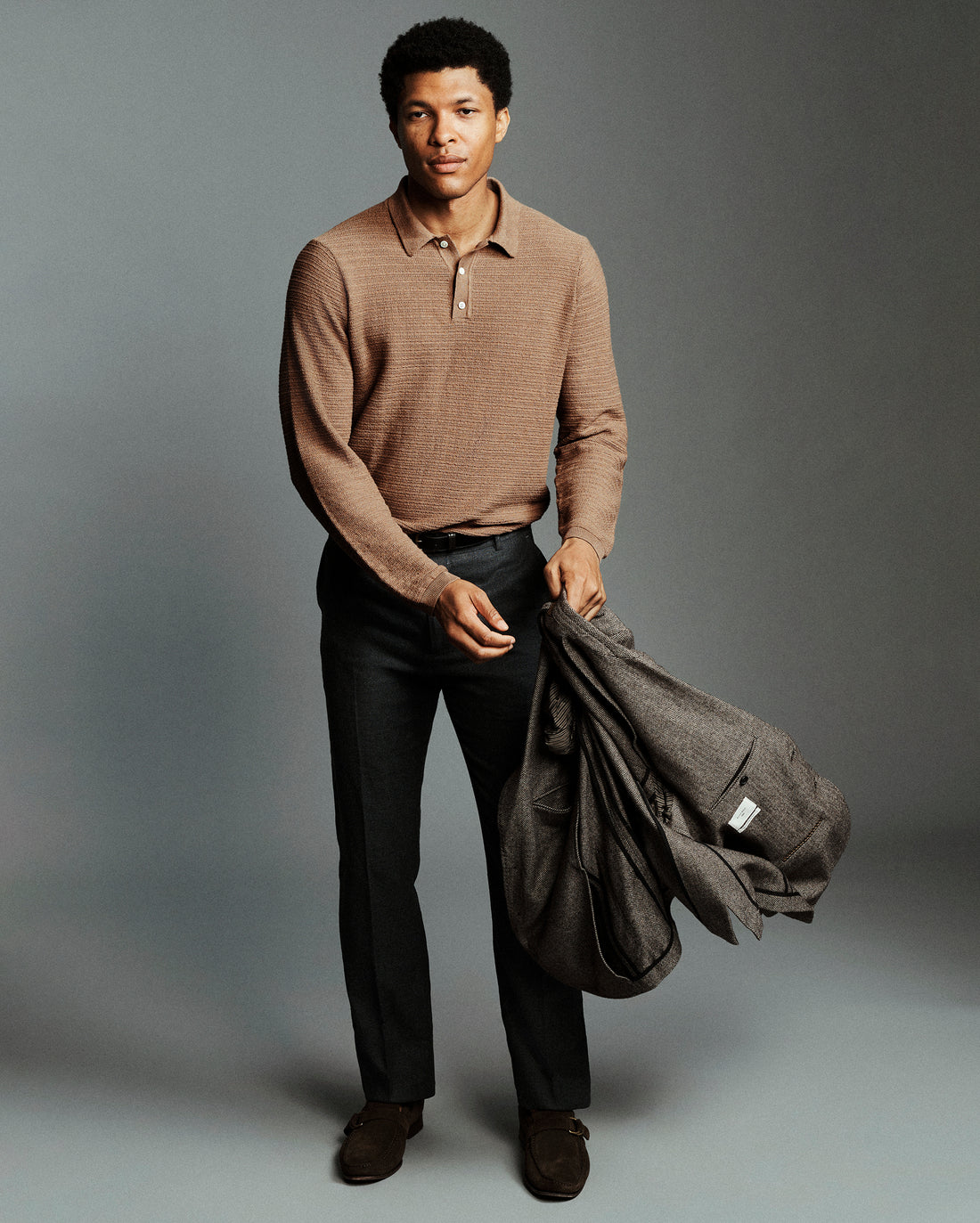 A man stands against a gray studio background wearing a tan ribbed knit polo shirt, dark trousers, and brown shoes, while holding a dark gray jacket.