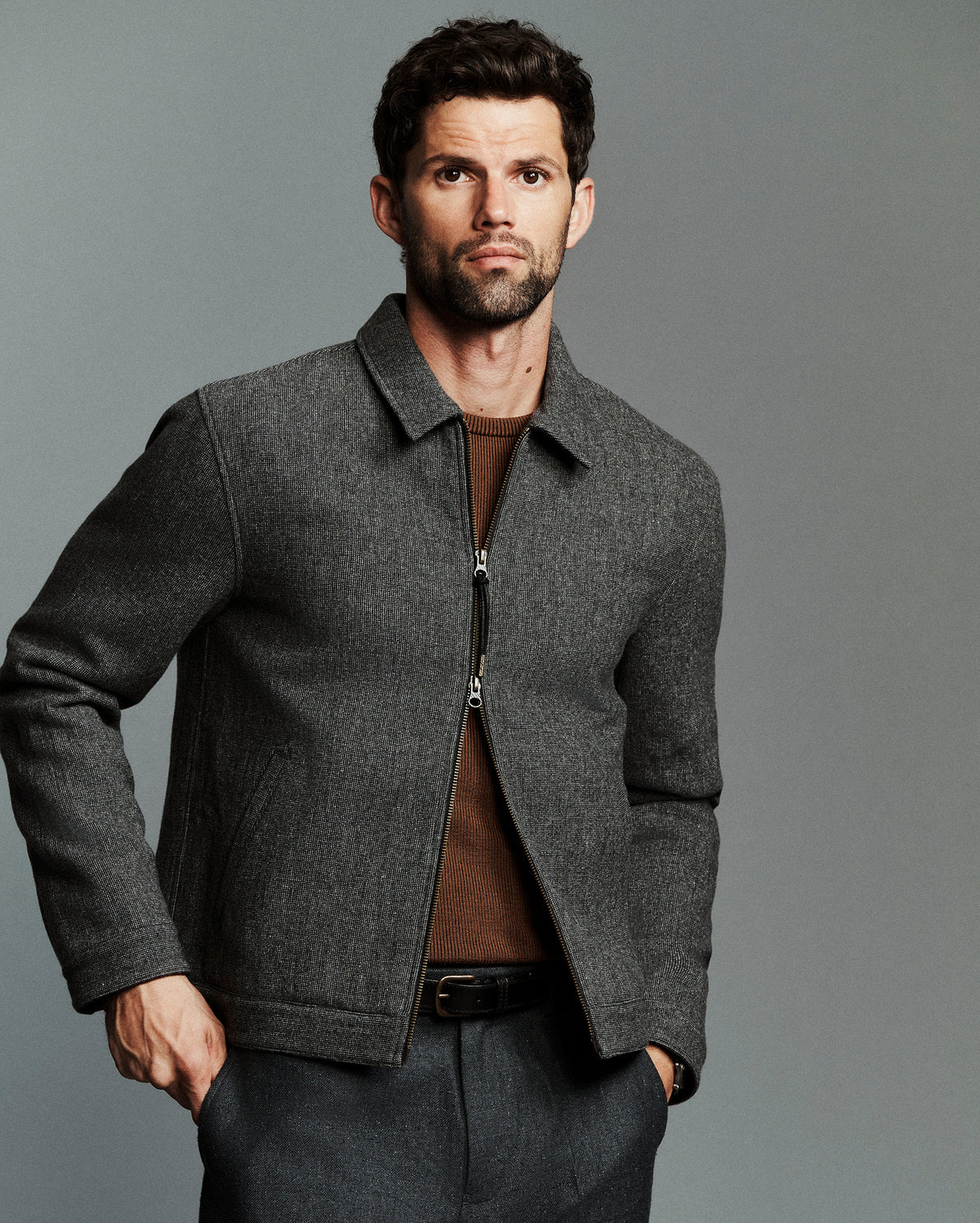 A man wearing a dark gray textured zip-up jacket over a brown shirt and gray trousers, standing against a solid gray studio background.