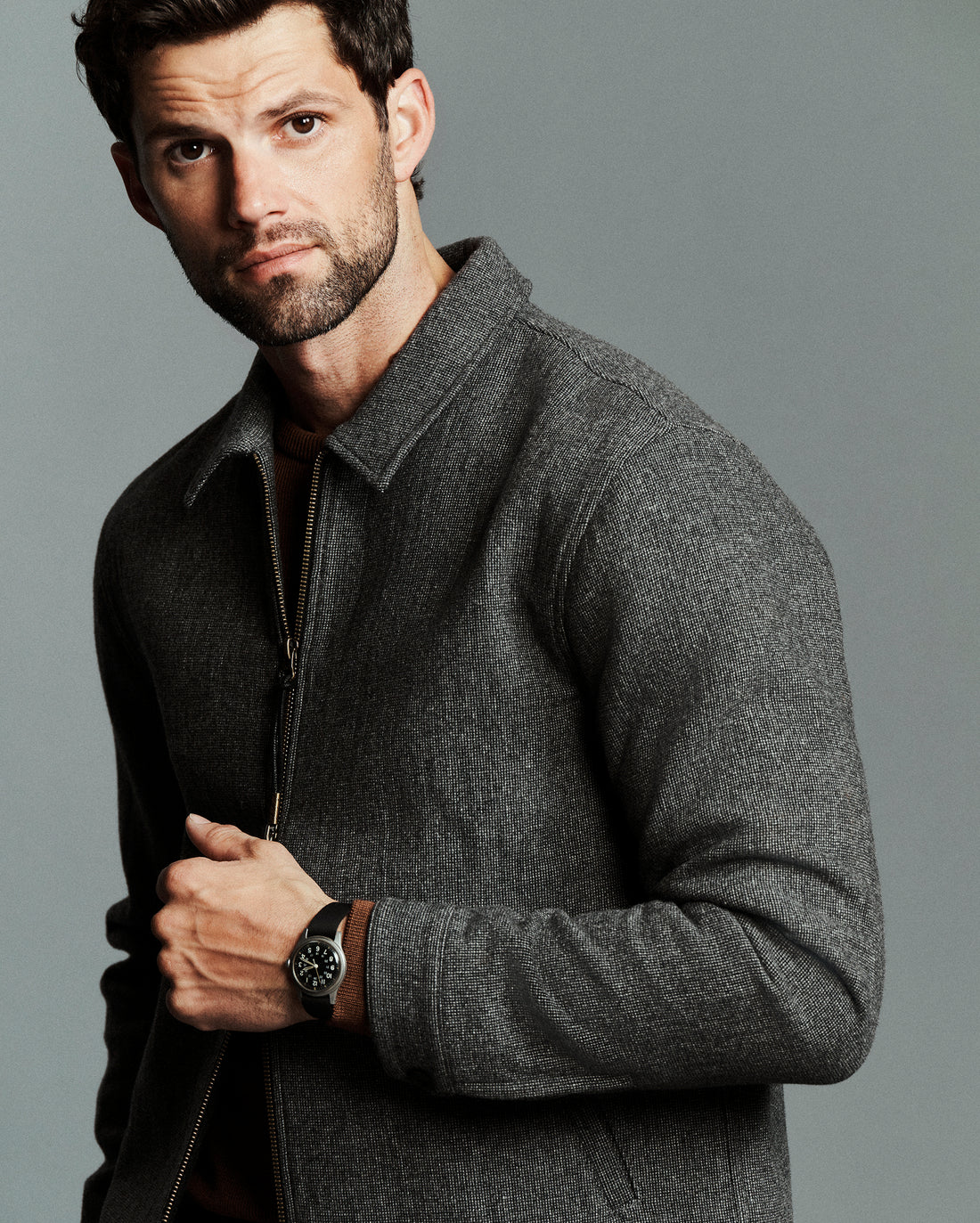 A man wearing a dark gray textured zip-up jacket and a black wristwatch, posed against a solid gray studio background.