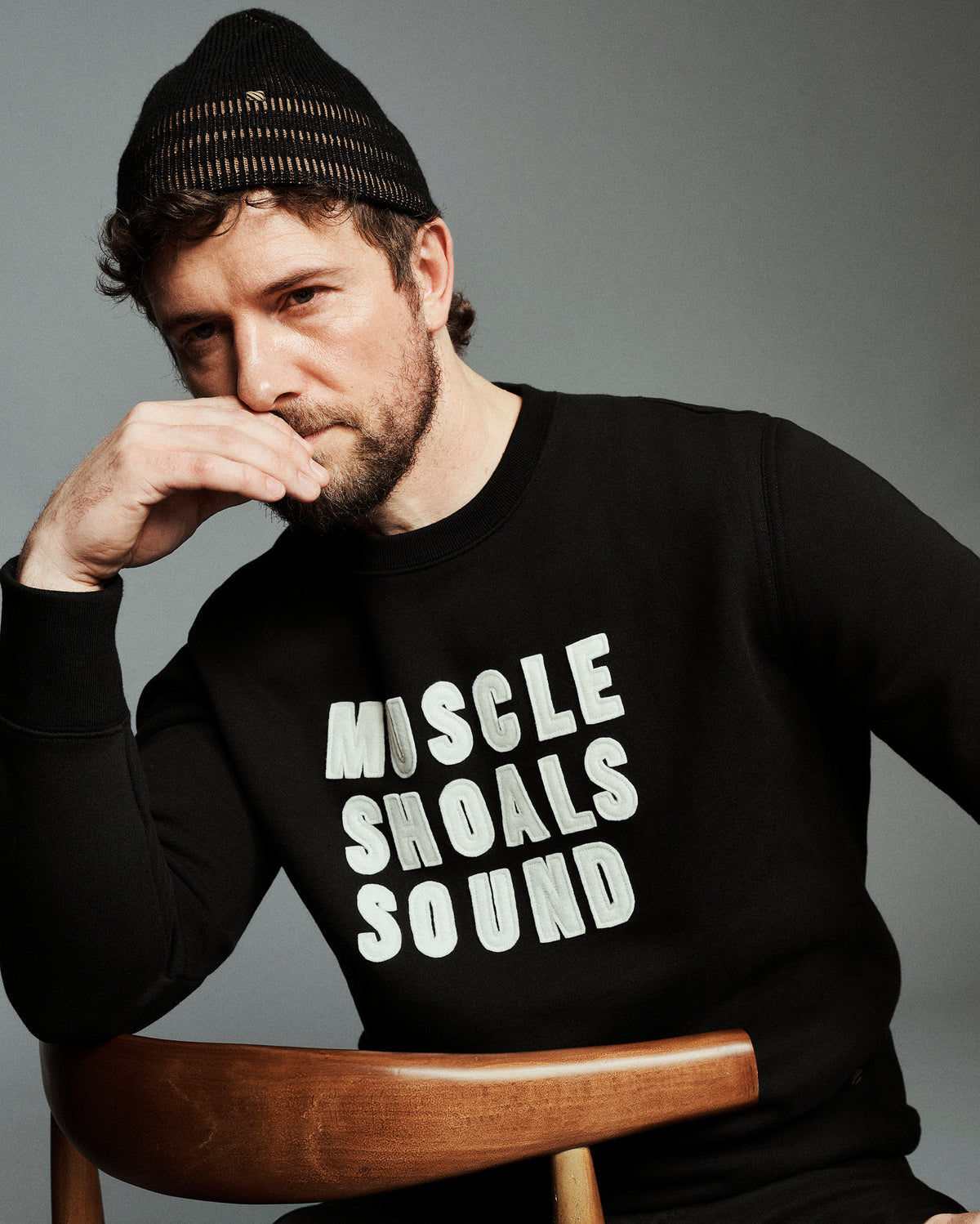 A man wearing a black knit beanie and a black long-sleeve sweatshirt with the text 'MUSCLE SHOALS SOUND' printed in white across the chest, seated on a wooden chair against a gray studio background.