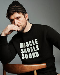 A man wearing a black knit beanie and a black long-sleeve sweatshirt with the text 'MUSCLE SHOALS SOUND' printed in white across the chest, seated on a wooden chair against a gray studio background.