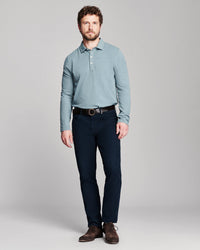 A man modeling a light blue long-sleeve knit polo shirt with dark navy pants and brown leather shoes, standing against a neutral gray studio background.