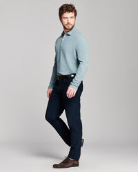 A man modeling a light blue long-sleeve knit polo shirt with dark navy trousers and brown leather shoes, standing against a neutral gray studio background.