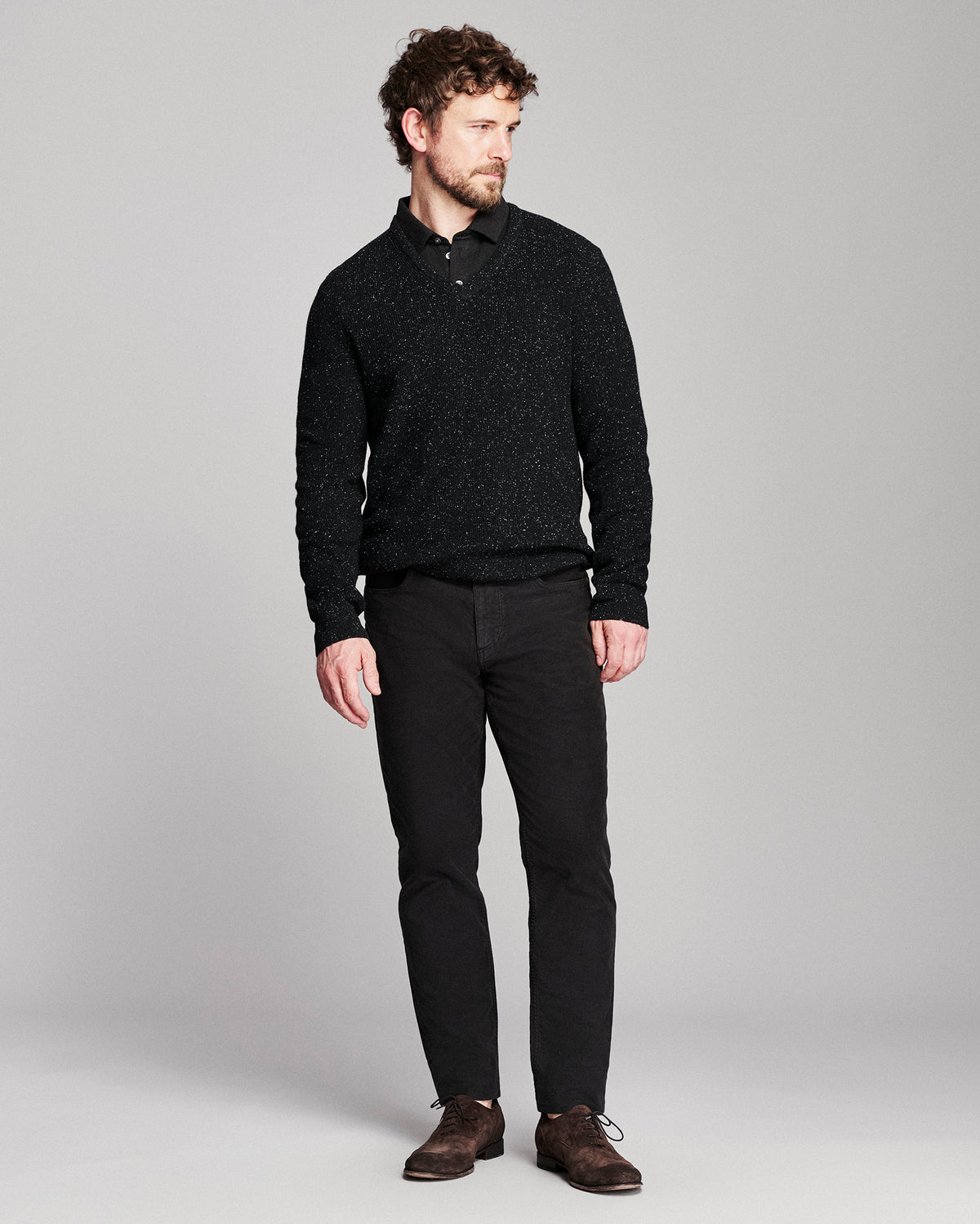 A man modeling a black textured knit polo shirt with black trousers and brown leather shoes, standing against a light gray studio background.