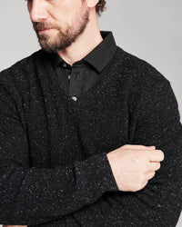 A close-up of a man wearing a black knit long-sleeve sweater with subtle white flecks over a black collared shirt, against a plain gray background.