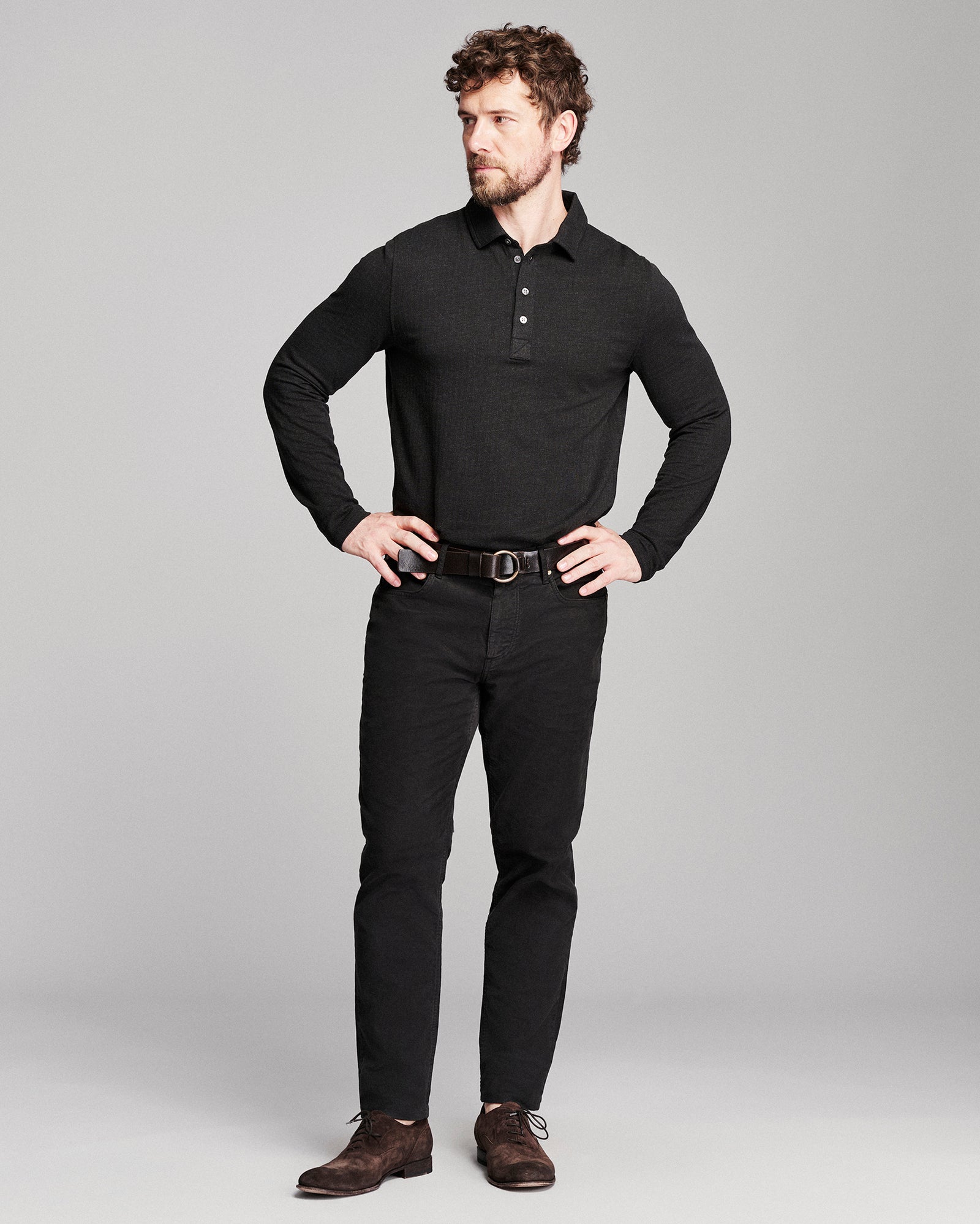 A man modeling a black long-sleeve polo shirt, black pants, a dark brown belt, and brown suede shoes against a light gray studio background.