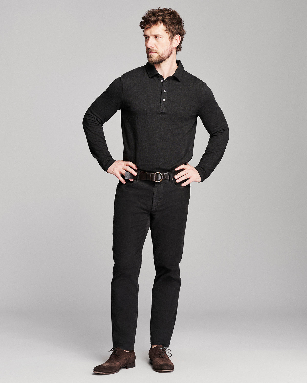 A man modeling a black long-sleeve polo shirt, black pants, a dark brown belt, and brown suede shoes against a light gray studio background.