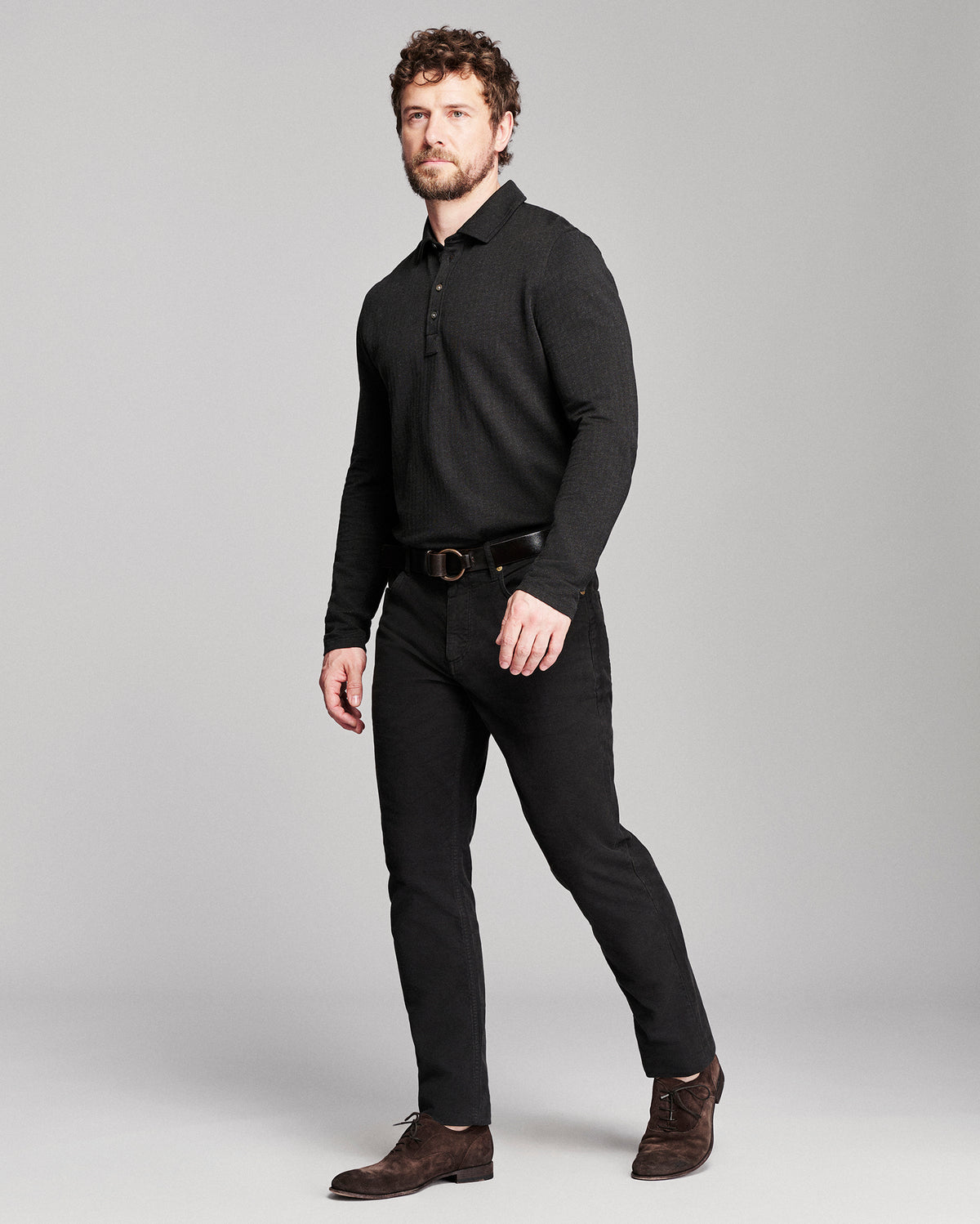 A man modeling a black long-sleeve knit polo shirt, black trousers, a black belt, and brown suede shoes against a solid gray studio background.