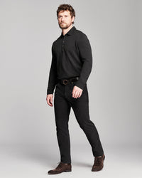 A man modeling a black long-sleeve knit polo shirt, black trousers, a black belt, and brown suede shoes against a solid gray studio background.