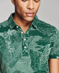 A close-up of a man wearing a short-sleeve polo shirt with a detailed green and teal dragon pattern, featuring a buttoned collar and chest pocket.