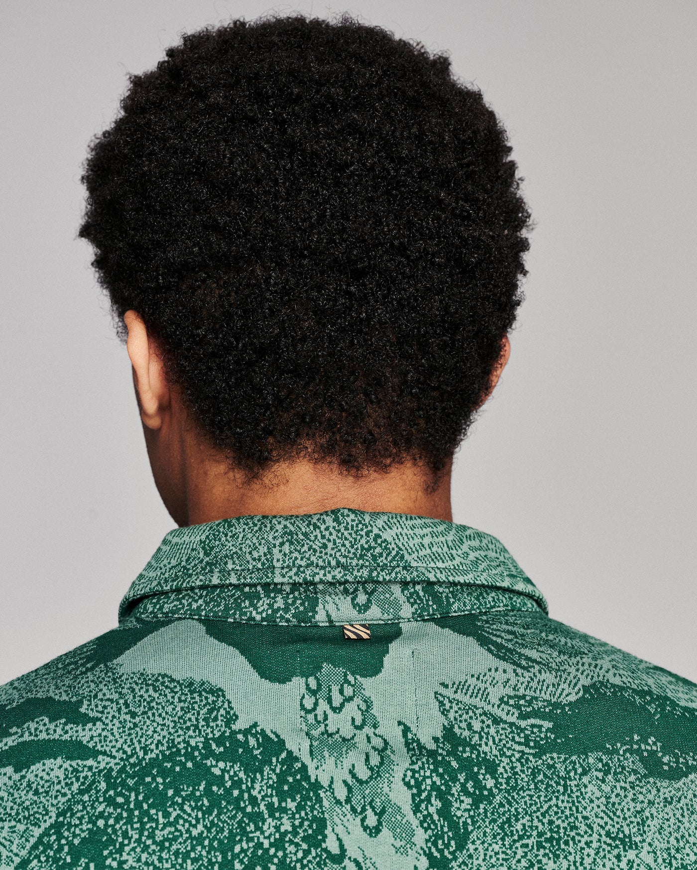 Back view of a person with curly black hair wearing a green patterned collared shirt against a light gray studio background.