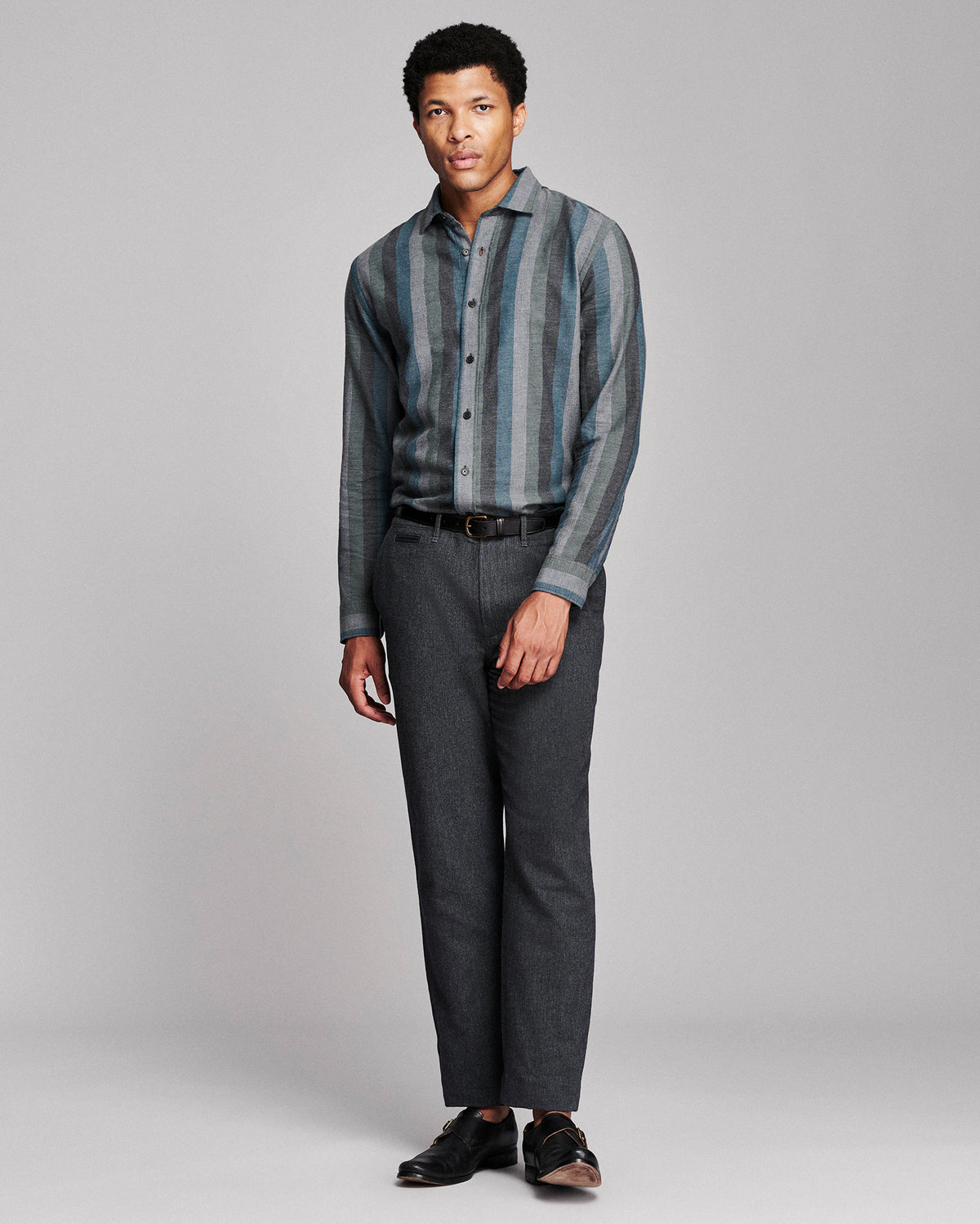 A man modeling a long-sleeve button-up shirt with vertical blue and gray stripes, paired with dark gray trousers and black shoes, against a light gray studio background.