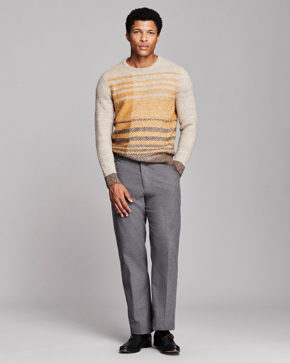 A man modeling a cream and mustard yellow striped knit sweater paired with gray trousers and black shoes, standing against a light gray studio background.