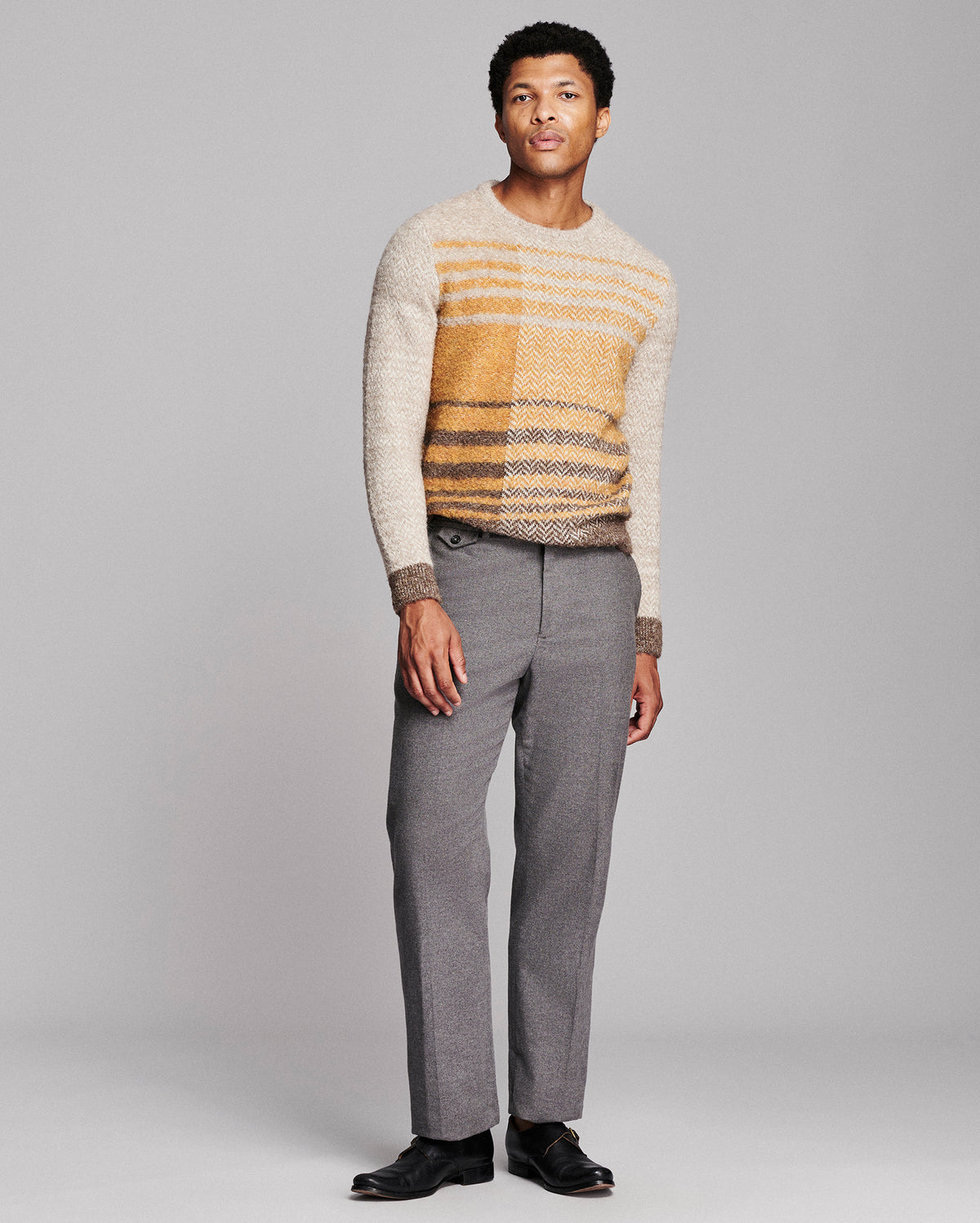 A man modeling a knit sweater with horizontal stripes in cream, mustard yellow, and brown, paired with gray trousers and black dress shoes, against a light gray studio background.