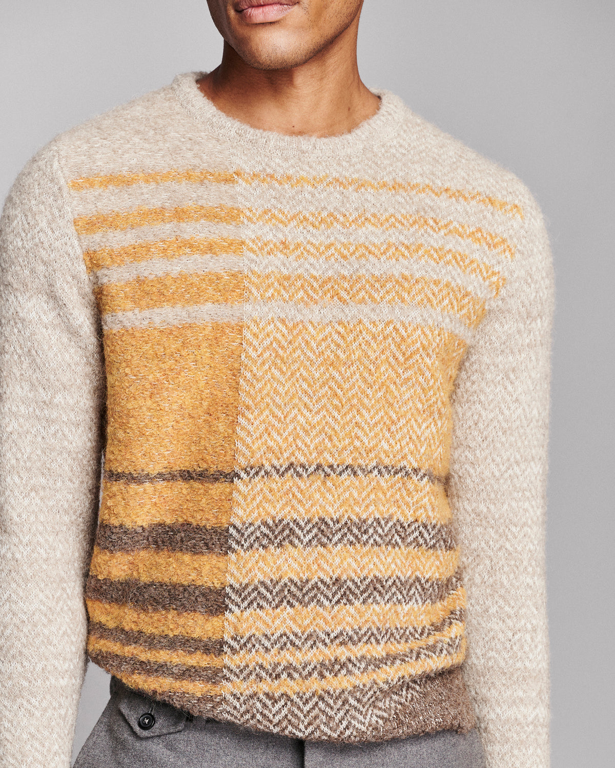 A close-up of a man wearing a fuzzy knit sweater with a patchwork design of yellow, beige, and brown horizontal stripes and herringbone patterns, paired with gray trousers against a solid gray background.