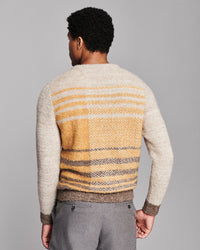 A man viewed from behind wearing a textured knit sweater with a cream body, yellow and brown horizontal stripes on the back, and gray trousers, set against a solid gray studio background.