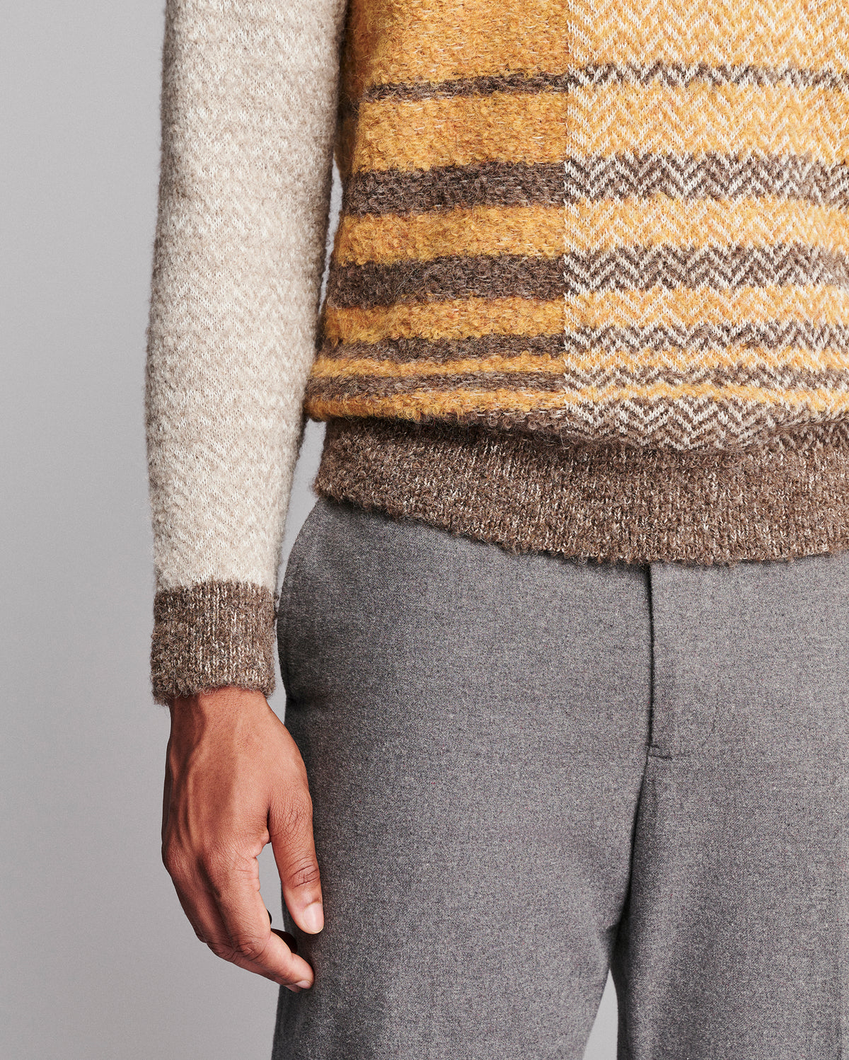 Close-up of a person wearing a fuzzy knit sweater with a cream sleeve, mustard yellow and brown stripes, and a brown ribbed hem, paired with gray trousers against a light gray background.