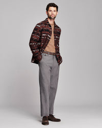 A man modeling a patterned button-down shirt with brown and red stripes, worn open over a tan sweater, paired with gray trousers and dark brown shoes, against a light gray studio background.