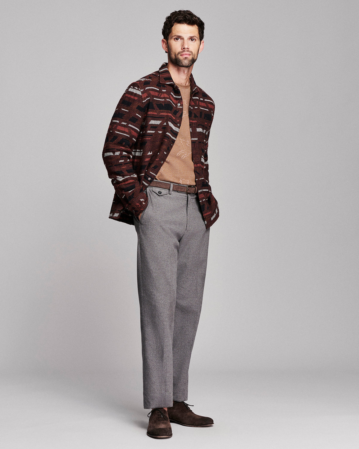 A man modeling a patterned button-down shirt with brown and red stripes, worn open over a tan sweater, paired with gray trousers and dark brown shoes, against a light gray studio background.