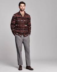 A man modeling a long-sleeve button-up shirt with a dark brown, red, and black abstract pattern, paired with gray trousers and brown shoes, against a solid gray studio background.