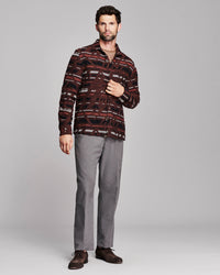 A man modeling a long-sleeve button-up shirt with a dark red, brown, and black geometric pattern, paired with gray trousers and brown shoes, against a light gray studio background.