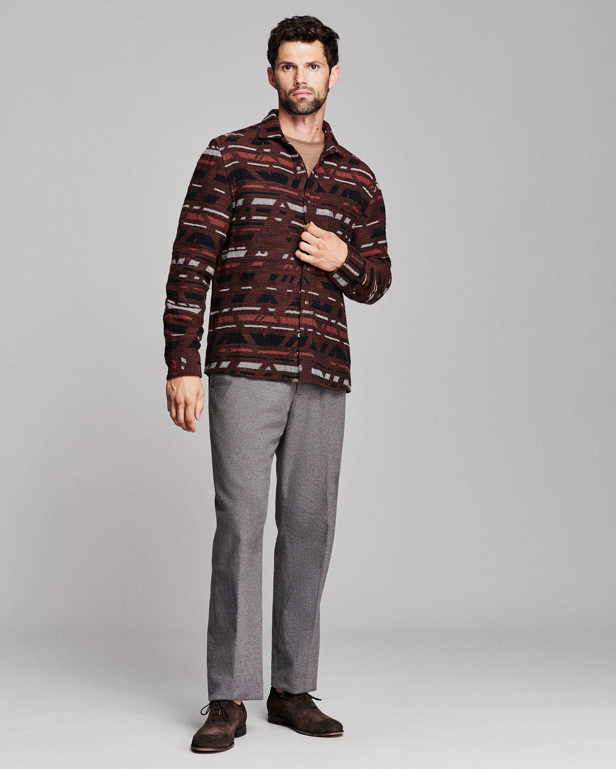 A man modeling a long-sleeve button-up shirt with a dark red, brown, and black geometric pattern, paired with gray trousers and brown shoes, against a light gray studio background.