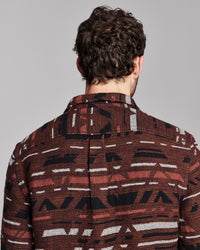 A man viewed from behind wearing a brown patterned button-down shirt with black and white geometric stripes, set against a solid gray studio background.