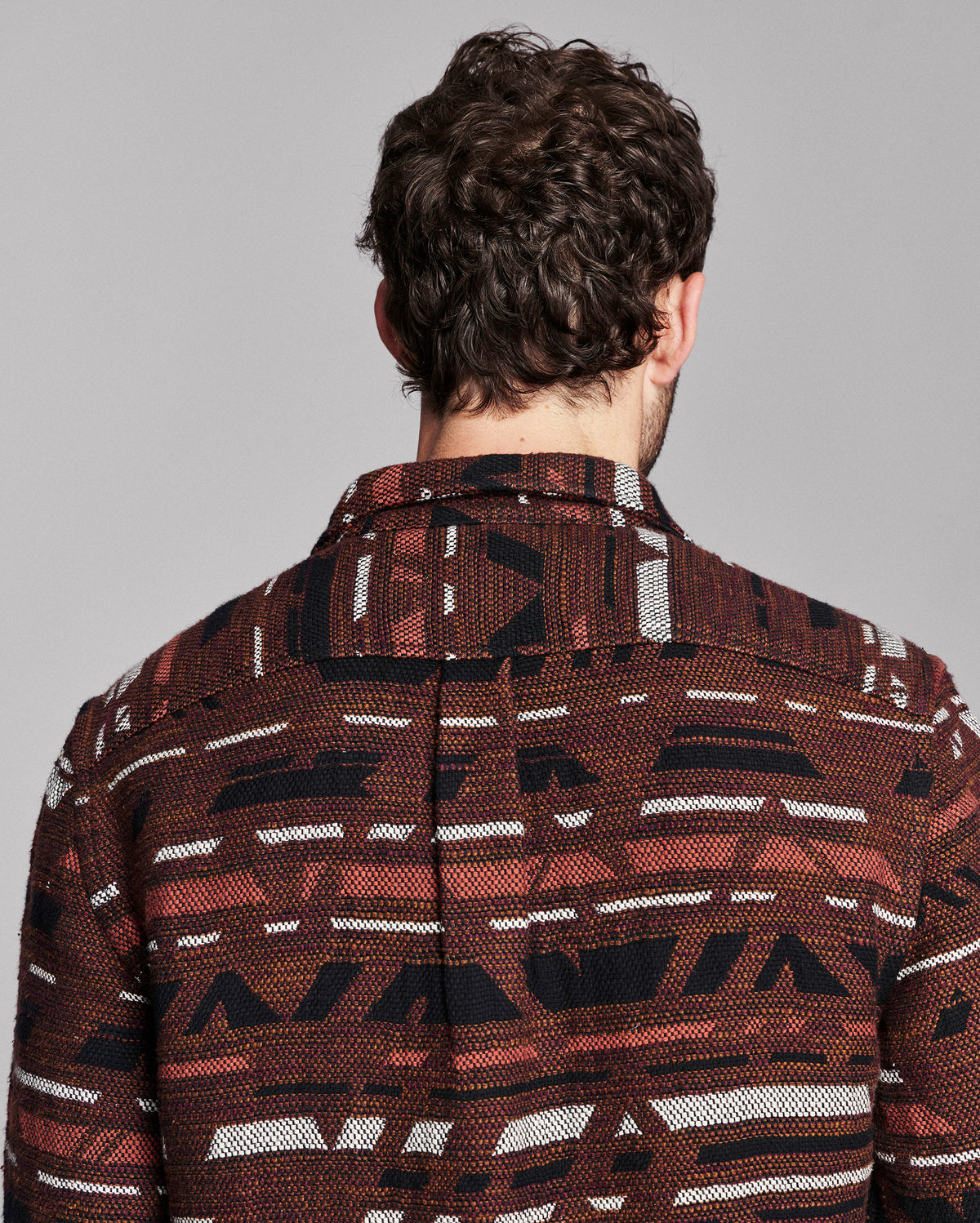 A man viewed from behind wearing a brown patterned button-down shirt with black and white geometric stripes, set against a solid gray studio background.