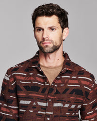 A man wearing a brown and black patterned button-down shirt over a beige undershirt, photographed against a solid gray background.