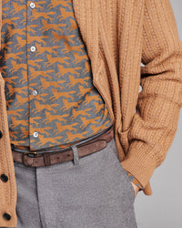 A close-up of a person wearing a tan ribbed knit cardigan over a gray button-up shirt with an orange fox pattern, paired with a brown belt and gray trousers.