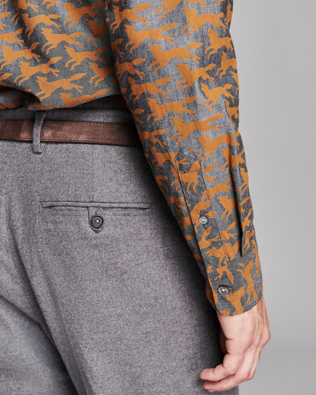 Close-up of a person wearing a long-sleeve button-up shirt with an orange and gray fox pattern, paired with gray trousers and a brown belt, shown from the waist down against a light gray studio background.