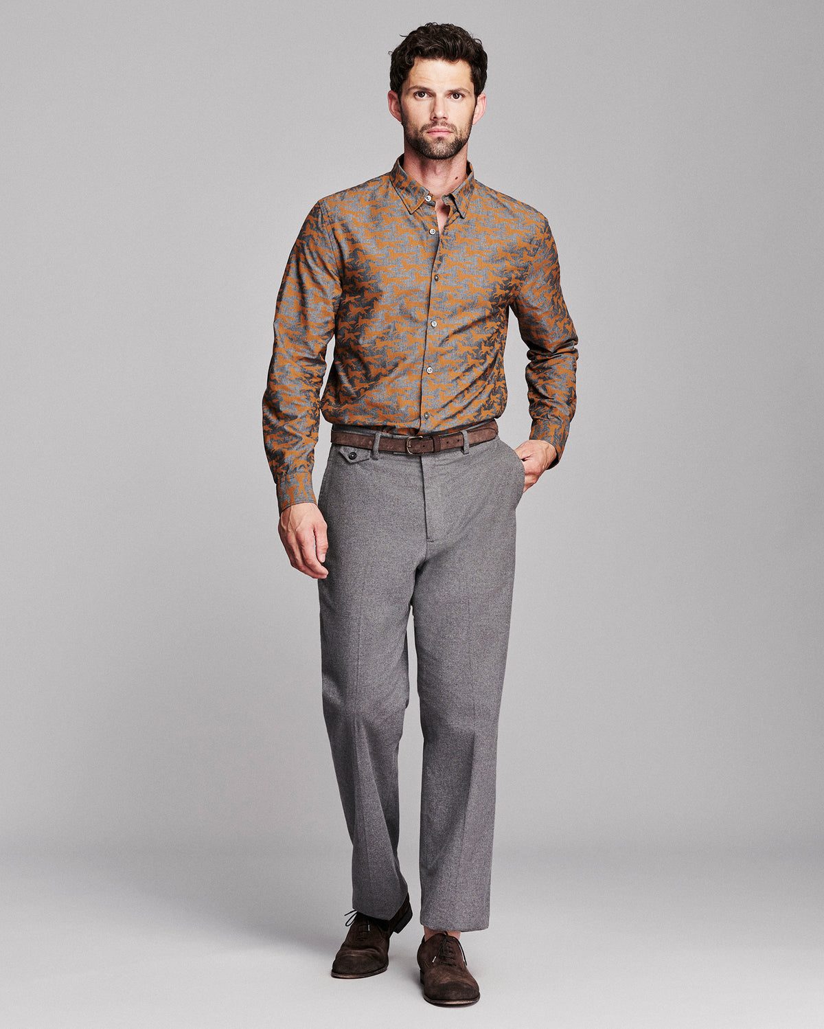 A man modeling a long-sleeve button-down shirt with a brown and orange fox pattern, paired with gray trousers and dark brown dress shoes, against a solid gray studio background.