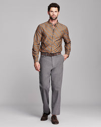 A man modeling a long-sleeve button-down shirt with a brown and orange fox pattern, paired with gray trousers and dark brown dress shoes, against a solid gray studio background.