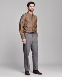 A man modeling a long-sleeve button-down shirt with a brown and orange fox pattern, paired with gray trousers and dark brown shoes, against a solid gray studio background.