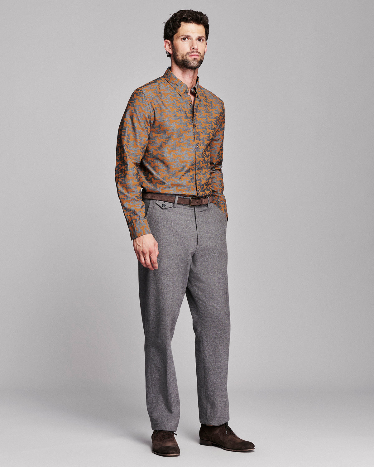 A man modeling a long-sleeve button-down shirt with a brown and orange fox pattern, paired with gray trousers and dark brown shoes, against a solid gray studio background.