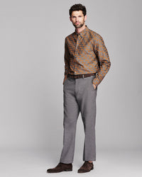 A man modeling a long-sleeve button-down shirt with an orange and brown fox pattern, paired with gray trousers and dark brown suede shoes, against a solid gray studio background.