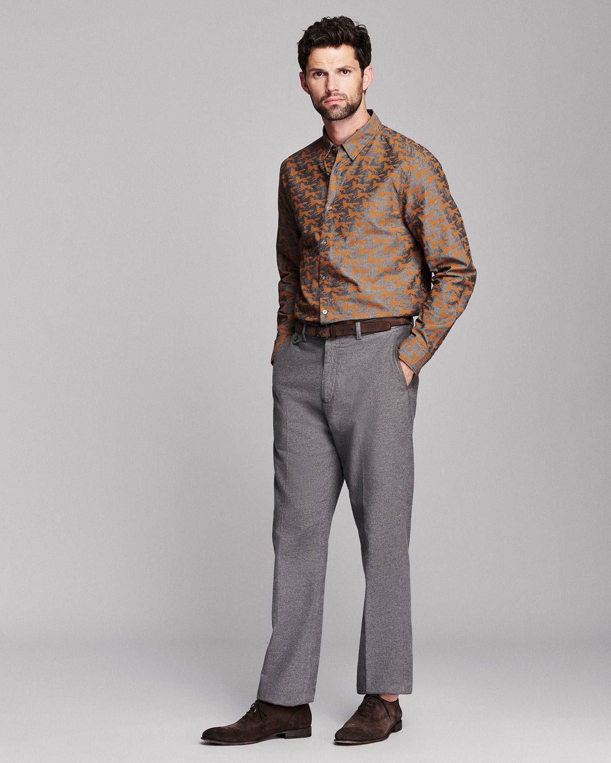 A man modeling a long-sleeve button-down shirt with an orange and brown fox pattern, paired with gray trousers and dark brown suede shoes, against a solid gray studio background.